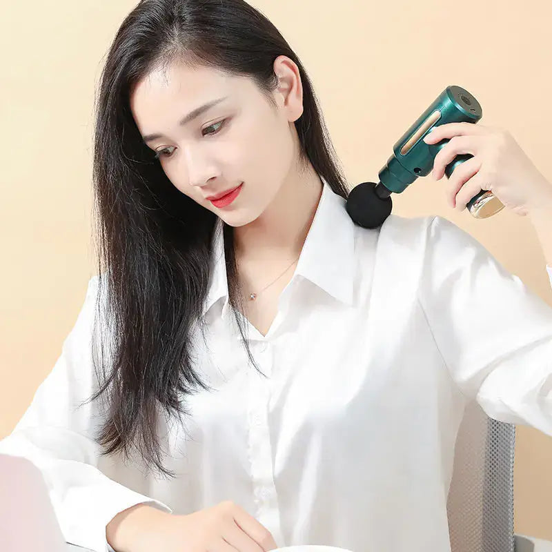 Woman using a professional fascia gun massager on her shoulder while working at a desk.
